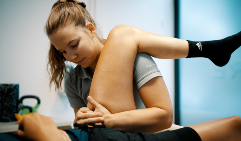 Physiotherapeutin umfasst bei einem liegenden Patienten den Unterschenkel nahe der Hüfte