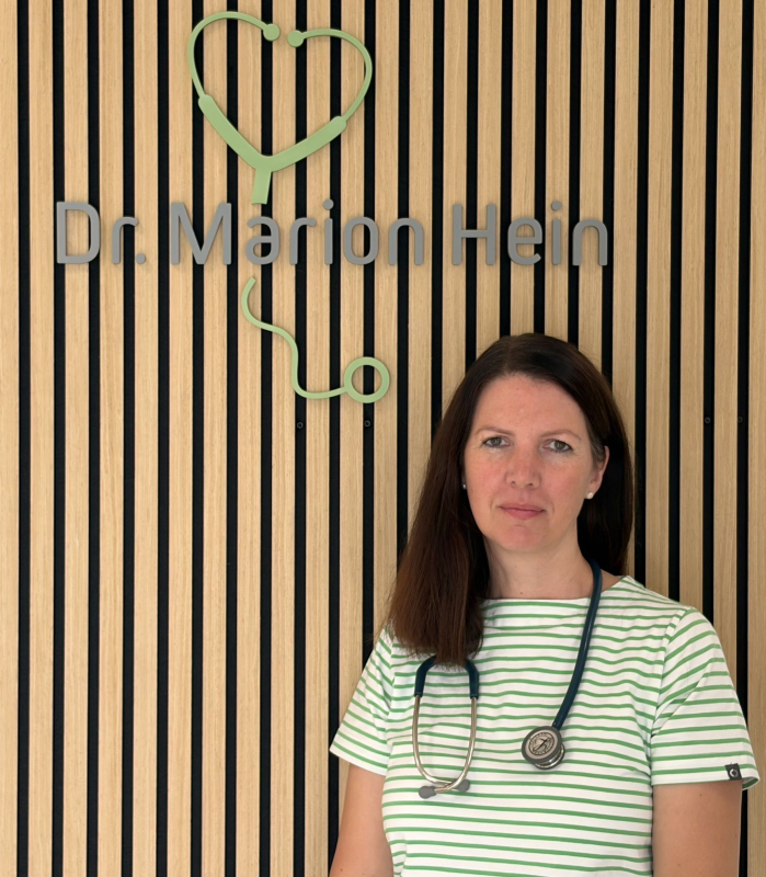 Profilbild der Fachärztin für Innere Medizin Dr. med. Marion Hein vor einer Wandpaneele mit Logo