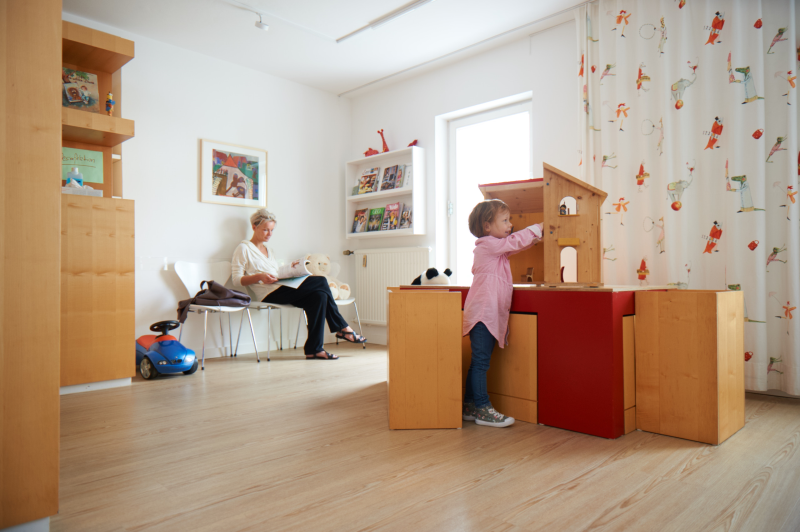 Kinderarztpraxis Ein Kind spielt an einem Spieltisch in einer Kinderarztpraxis, während eine Mutter im Hintergrund liest.