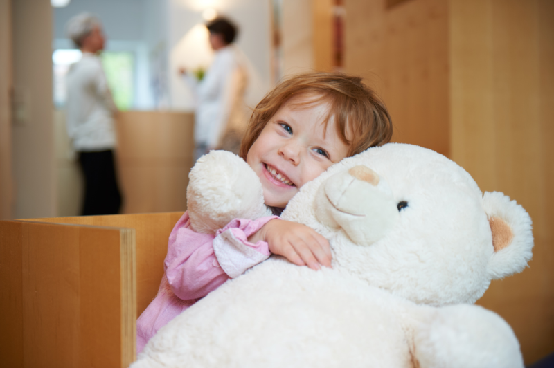Kind mit Teddy Ein kleines Mädchen umarmt einen großen Teddybär und lächelt fröhlich in einer Arztpraxis.
